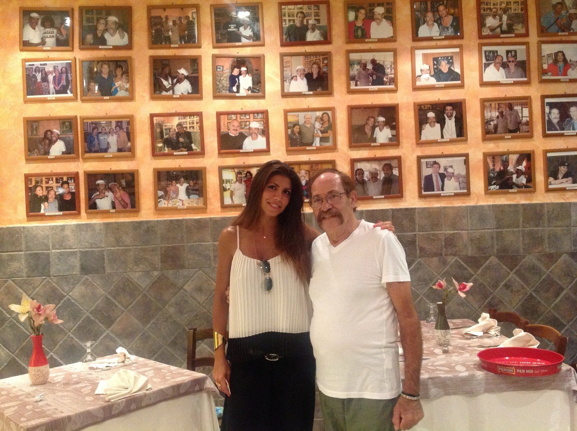 Due persone in posa in un ristorante con una parete di foto incorniciate alle loro spalle.