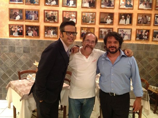 Tre persone sorridenti posano insieme in un ristorante, davanti a una parete ricoperta di fotografie incorniciate.