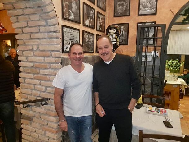 Due uomini in piedi insieme in un accogliente caffè con pareti in mattoni, sorridono alla macchina fotografica.