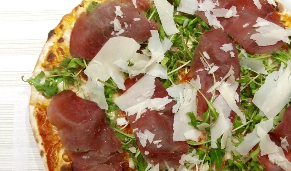 Primo piano di una pizza condita con rucola, prosciutto crudo e scaglie di parmigiano.