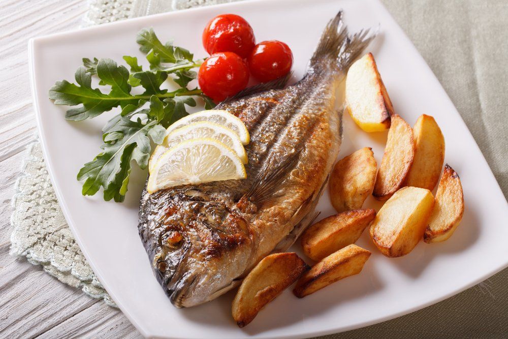 Pesce grigliato con limone, prezzemolo, pomodori e spicchi di patate su un piatto bianco.