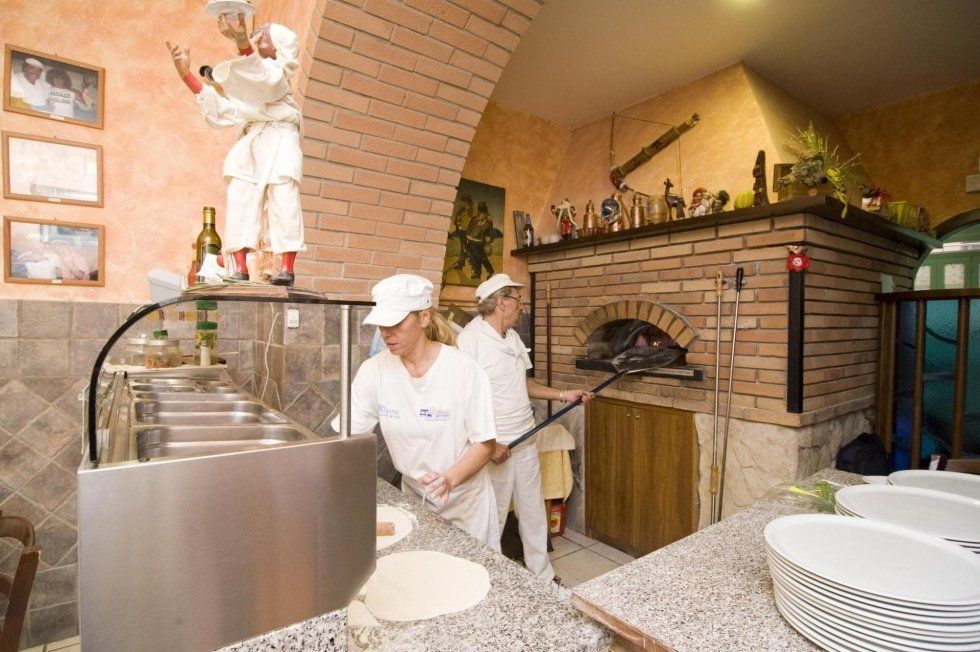 Due panettieri in uniforme bianca lavorano accanto a un forno a legna per la pizza nella cucina rustica di un ristorante.