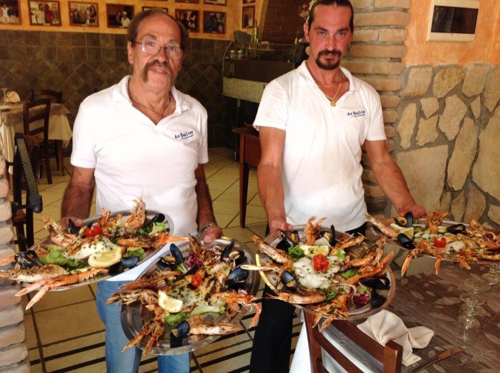 Due chef in camicia bianca mostrano diversi piatti di frutti di mare colorati su un tavolo di un ristorante.