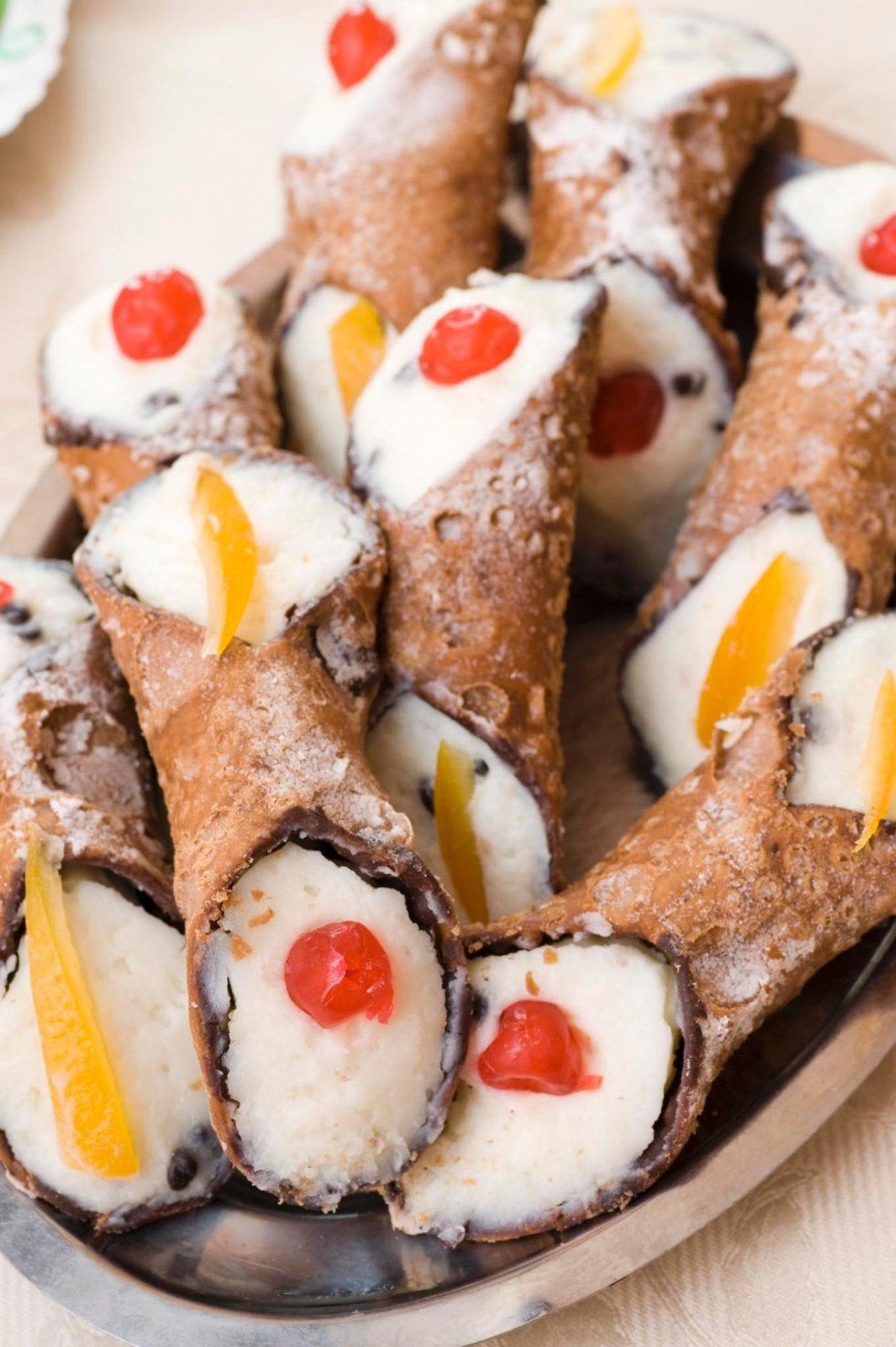 Vassoio di cannoli spolverati di zucchero a velo, ripieni di crema bianca e guarniti con ciliegie rosse e gialle.