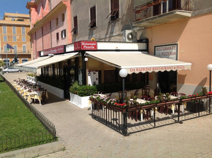 Caffè all'aperto con tenda da sole rossa e patio fiorito accanto a una strada acciottolata.