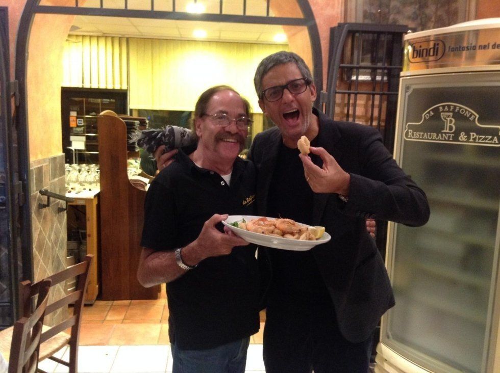 Due uomini sorridenti in un ristorante, uno con in mano un piatto di cibo e l'altro che ne mangia un boccone.