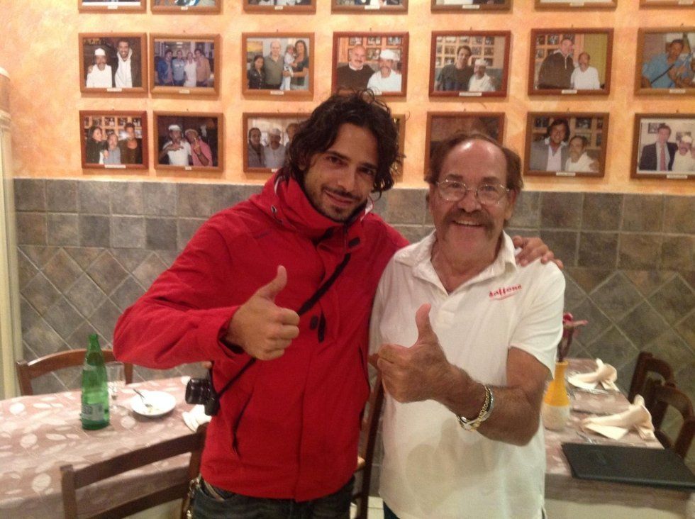 Due uomini sorridenti che fanno il segno del pollice in su in un ristorante, uno con una giacca rossa e l'altro con una camicia bianca.