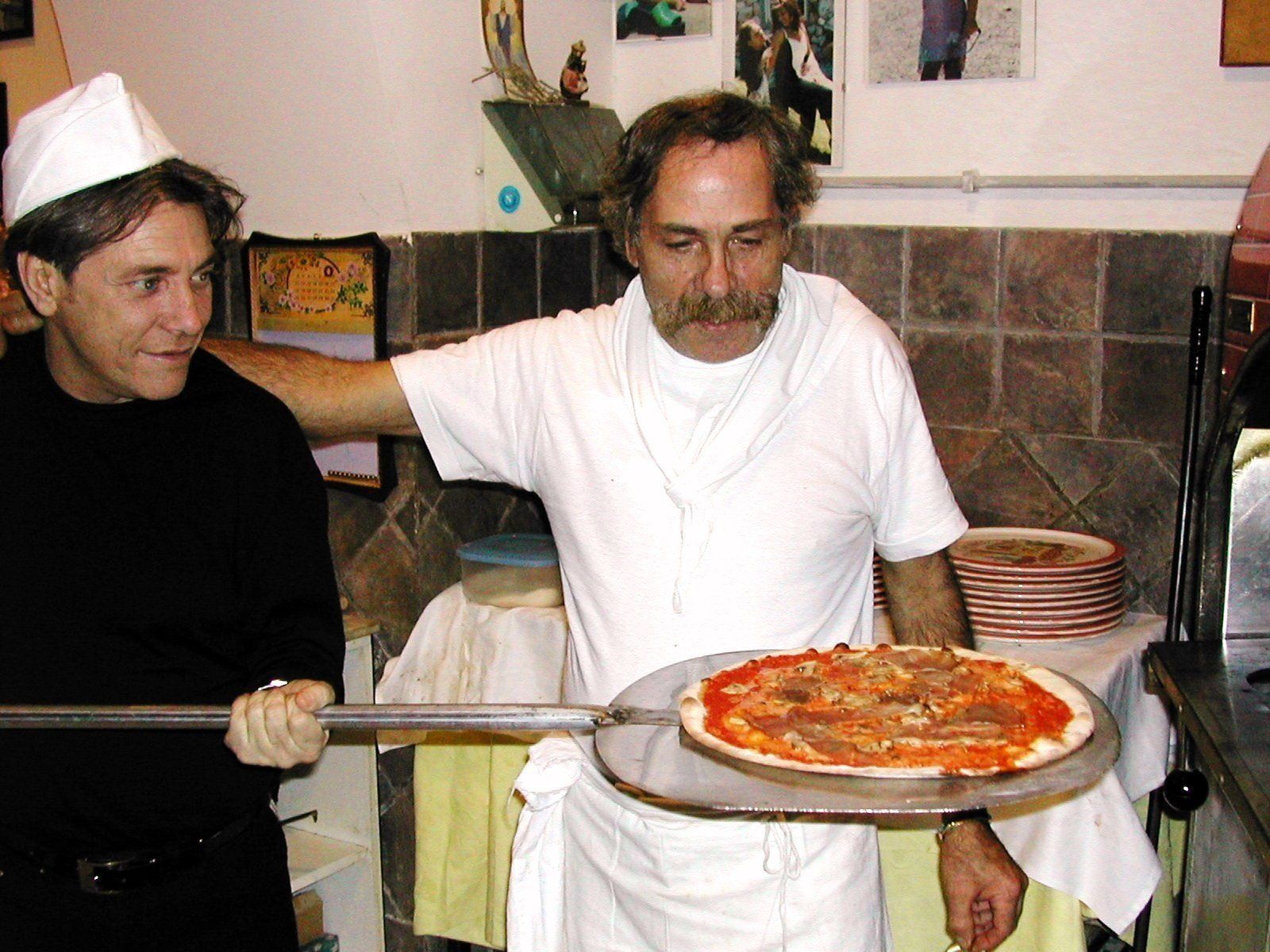 In una cucina, due uomini tengono in mano una pizza su un vassoio: uno indossa una camicia e un grembiule bianchi, l'altro abiti neri.