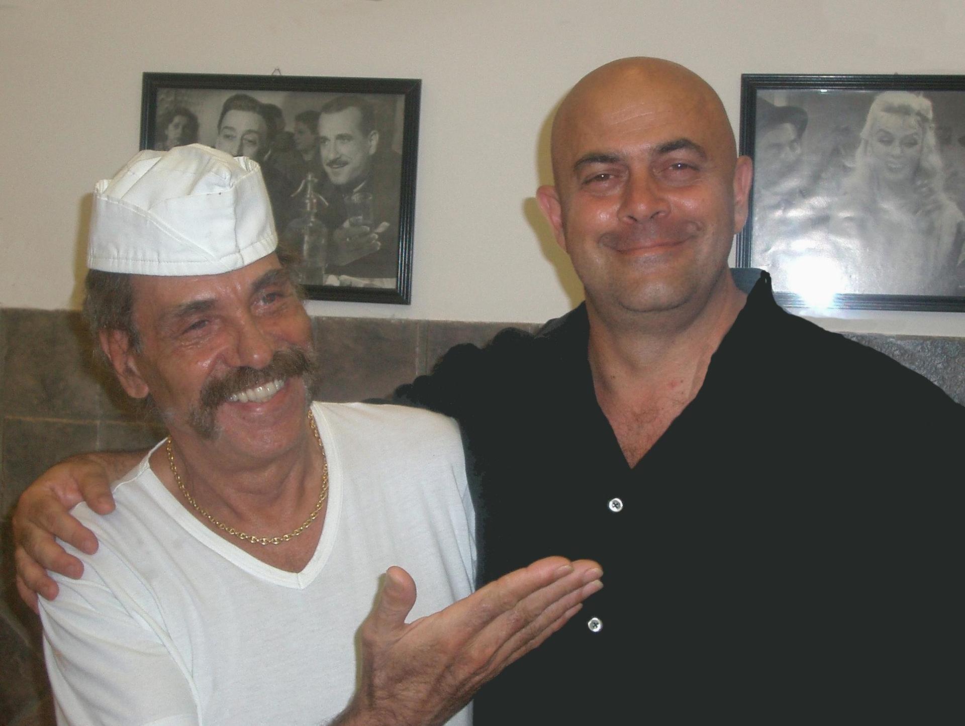 Due uomini sorridenti in posa al chiuso, uno con berretto e camicia bianchi, l'altro con una camicia nera e un braccio intorno a lui