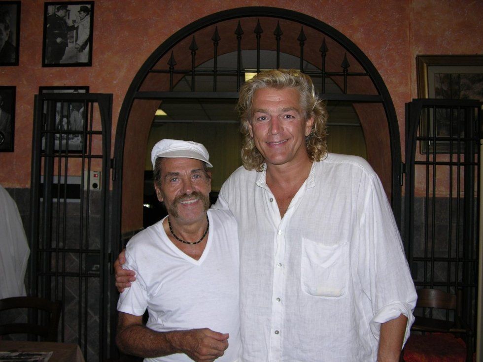 Due uomini in posa all'interno, uno con camicia e berretto bianchi, in piedi davanti a un portale ad arco.