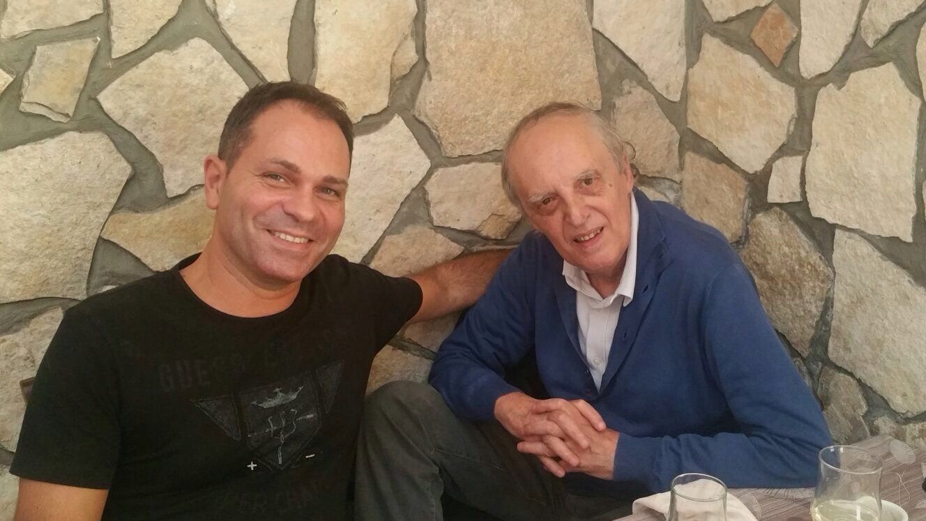 Due uomini sorridenti siedono a un tavolo davanti a un muro di pietra, posando insieme.