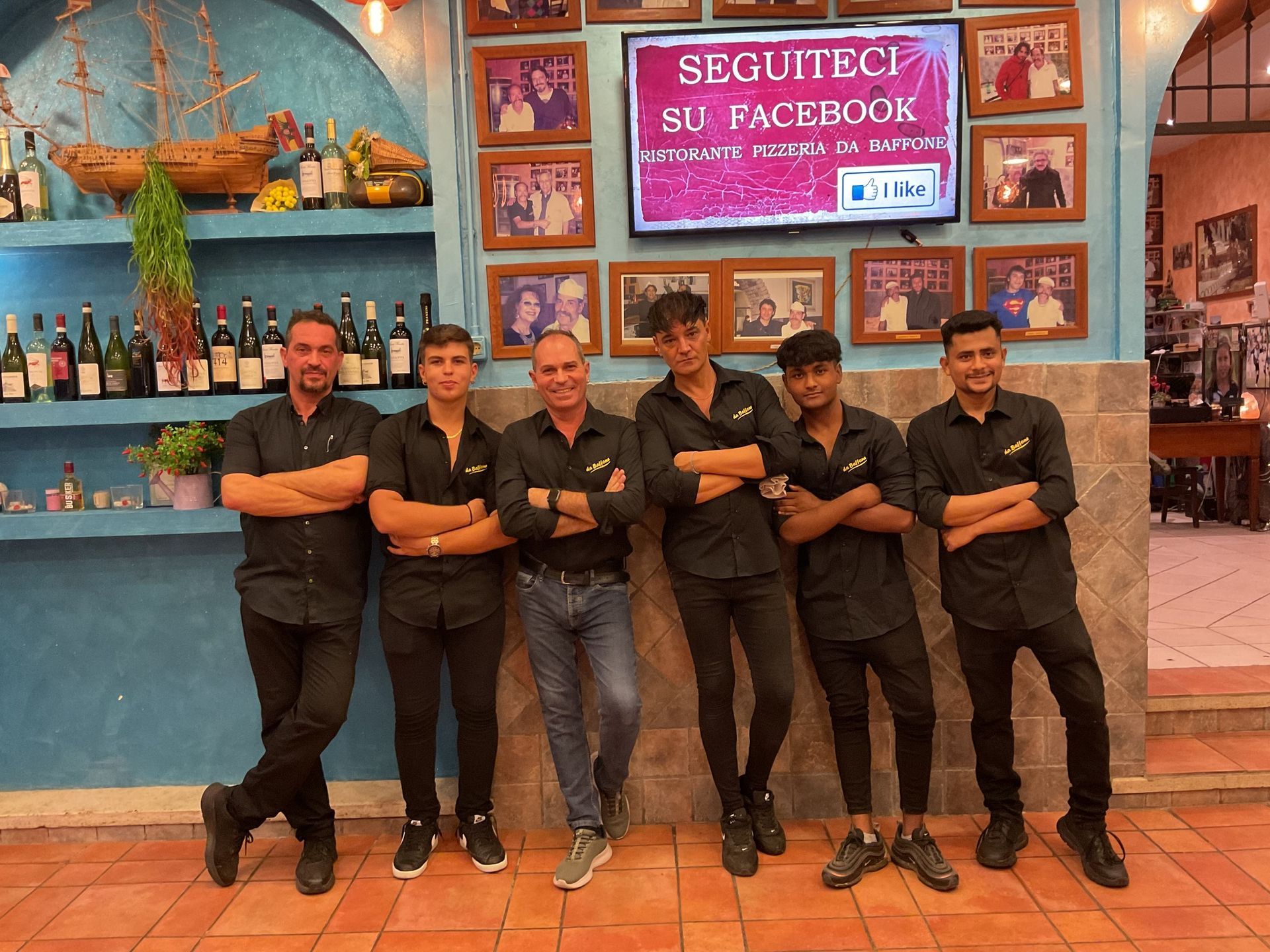 Cinque dipendenti in uniforme nera sono in piedi con le braccia incrociate davanti a un colorato bancone di un bar.