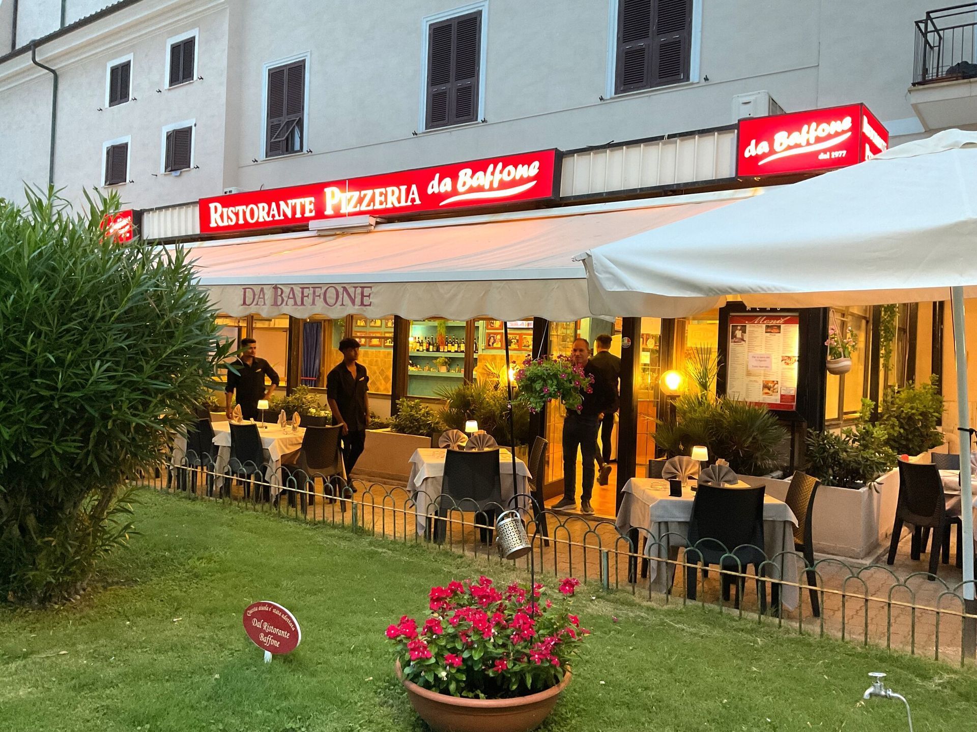 Terrazza esterna del ristorante con insegna rossa, tenda da sole bianca, tavoli e vasi di fiori al crepuscolo.