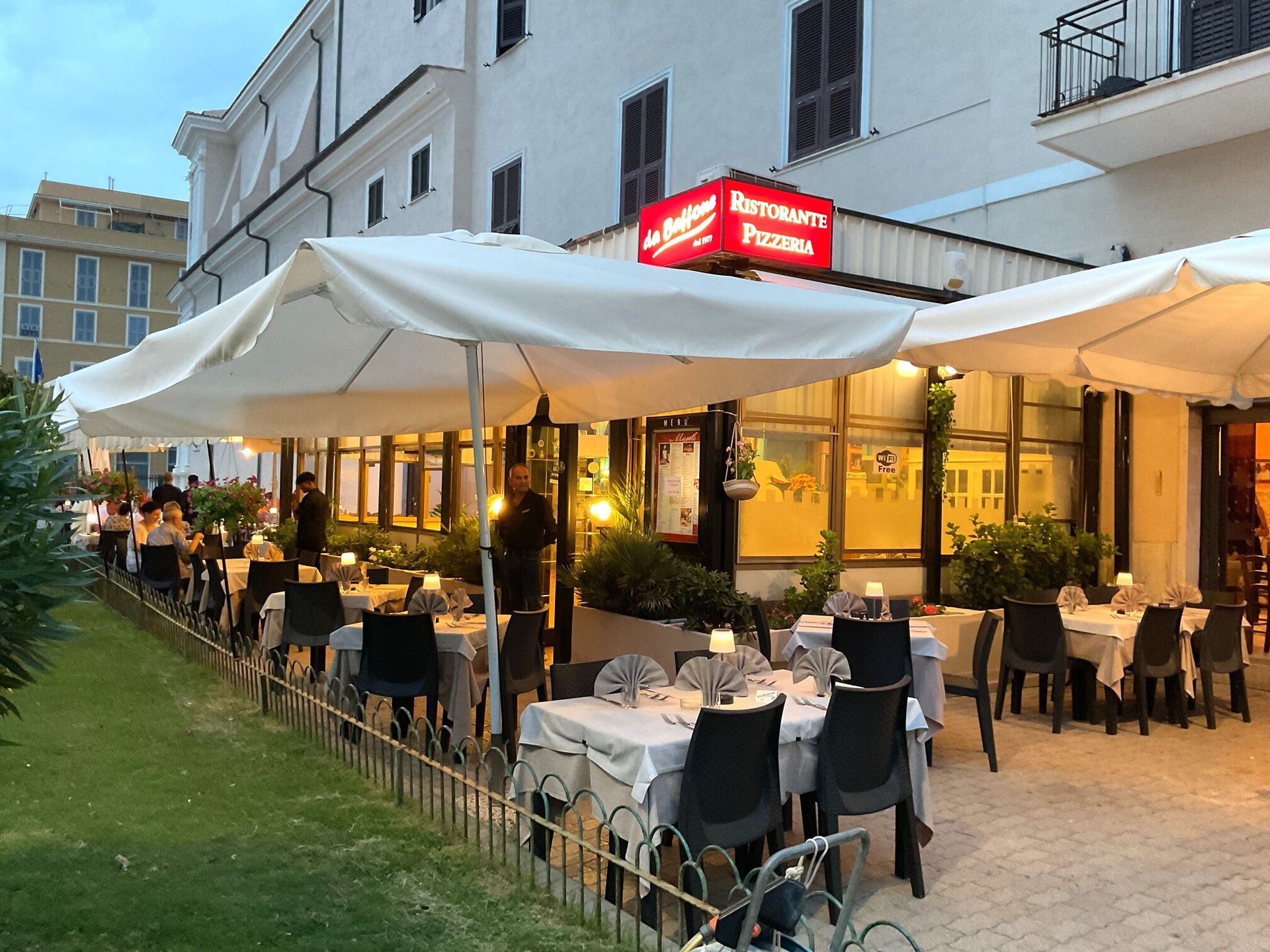 Terrazza esterna del ristorante con ombrelloni bianchi, tavoli illuminati da luci calde e clienti che pranzano lungo un sentiero erboso.