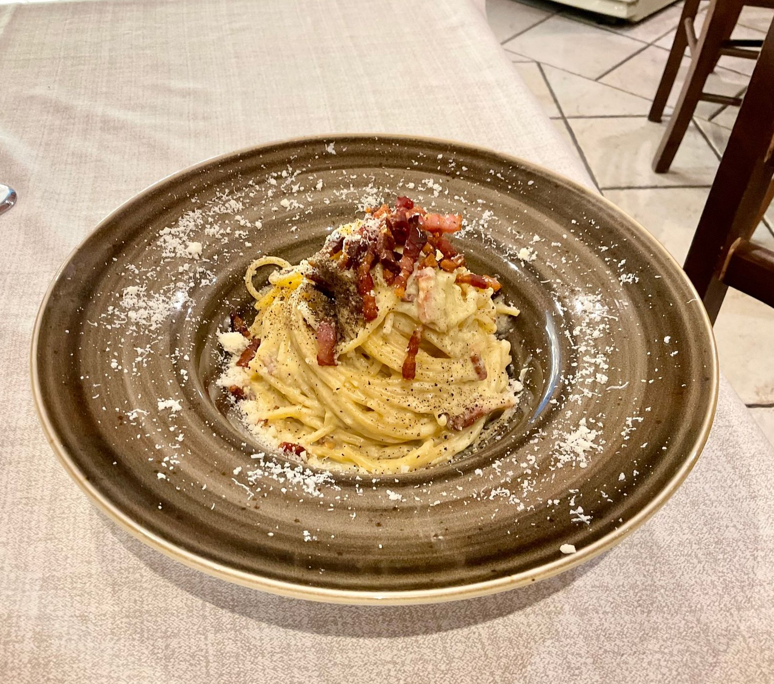 Un piatto di pasta in una ciotola marrone, guarnito con formaggio grattugiato e pezzetti di pancetta, su un tavolo di un ristorante.