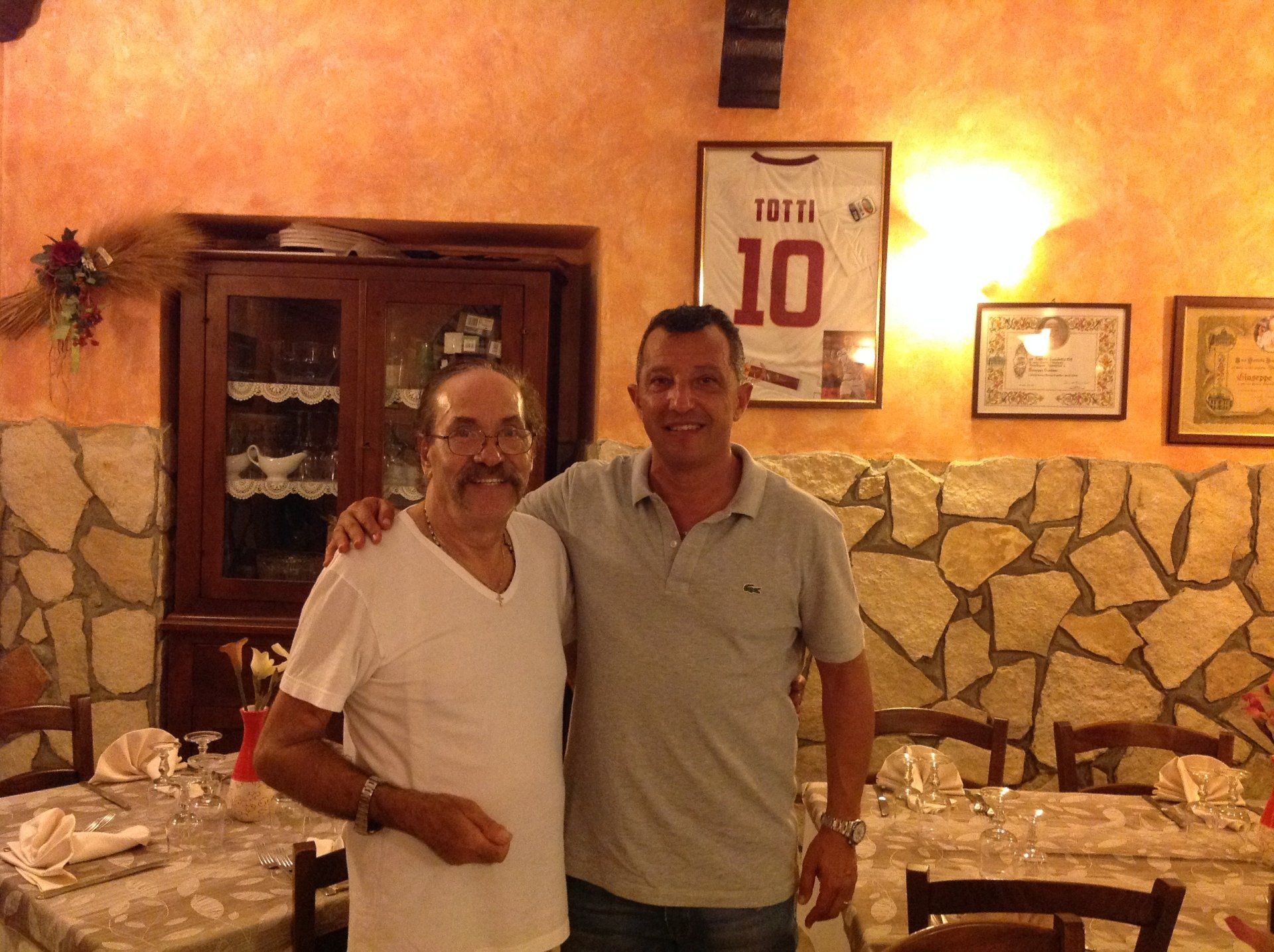 Due uomini sorridenti posano insieme in un ristorante illuminato da una luce calda, con pareti in pietra e decorazioni incorniciate.