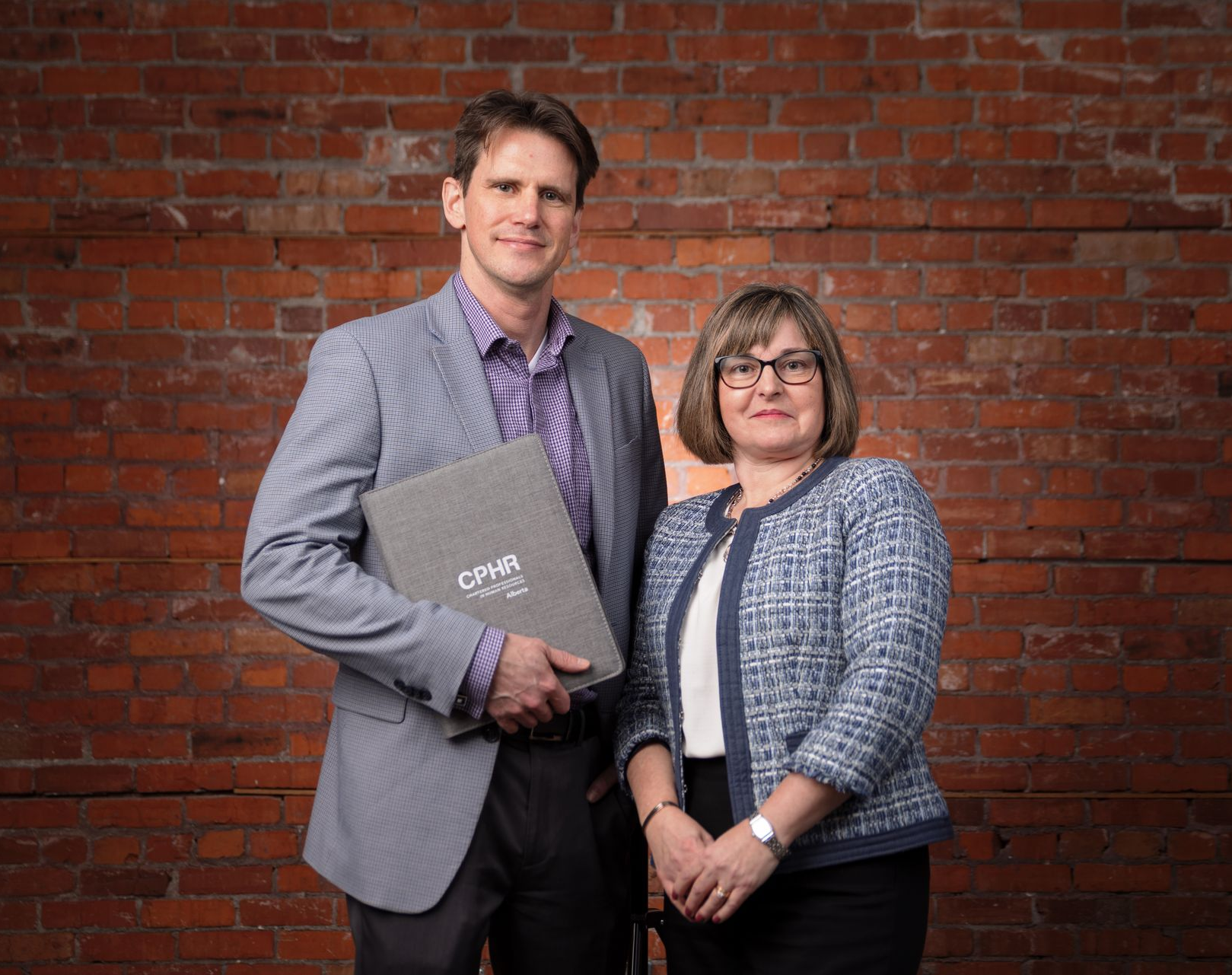 Man and woman standing in front of a brick wall, the man holds a folder, both smiling.