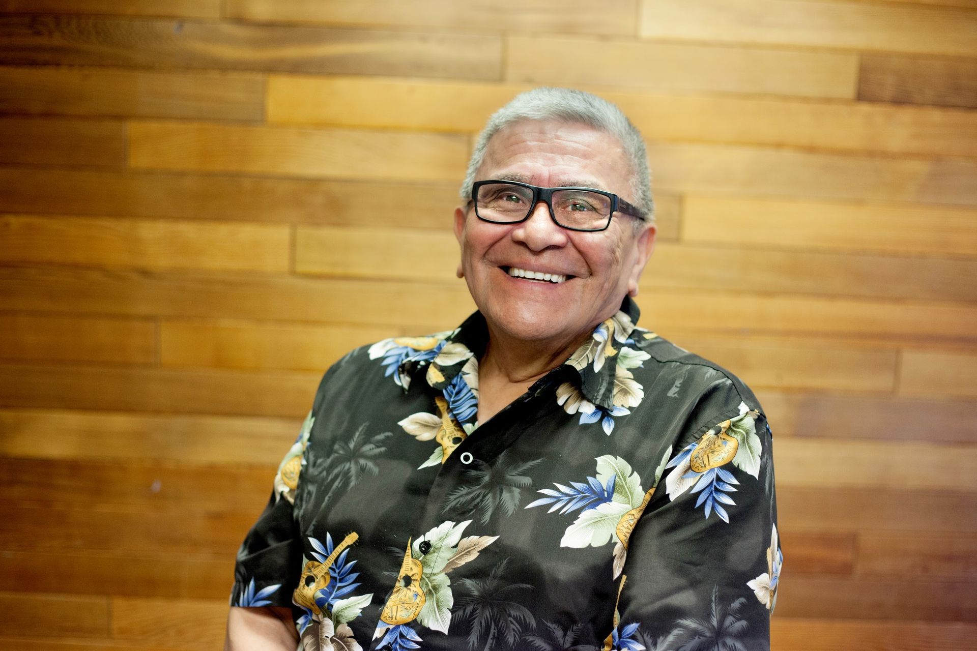 A man wearing glasses and a hawaiian shirt is smiling in front of a wooden wall.