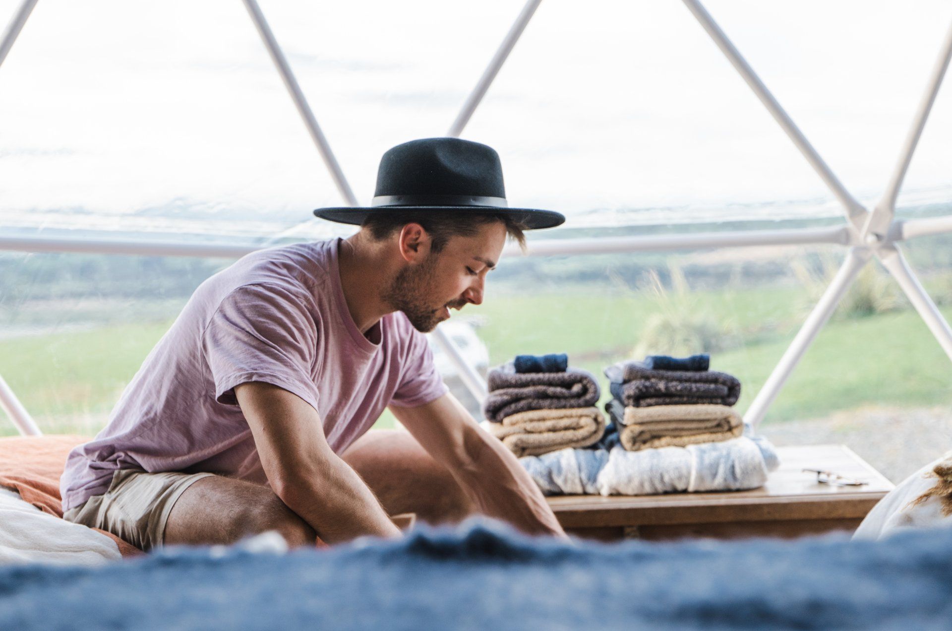 A man in a hat is sitting on a bed next to a stack of towels.
