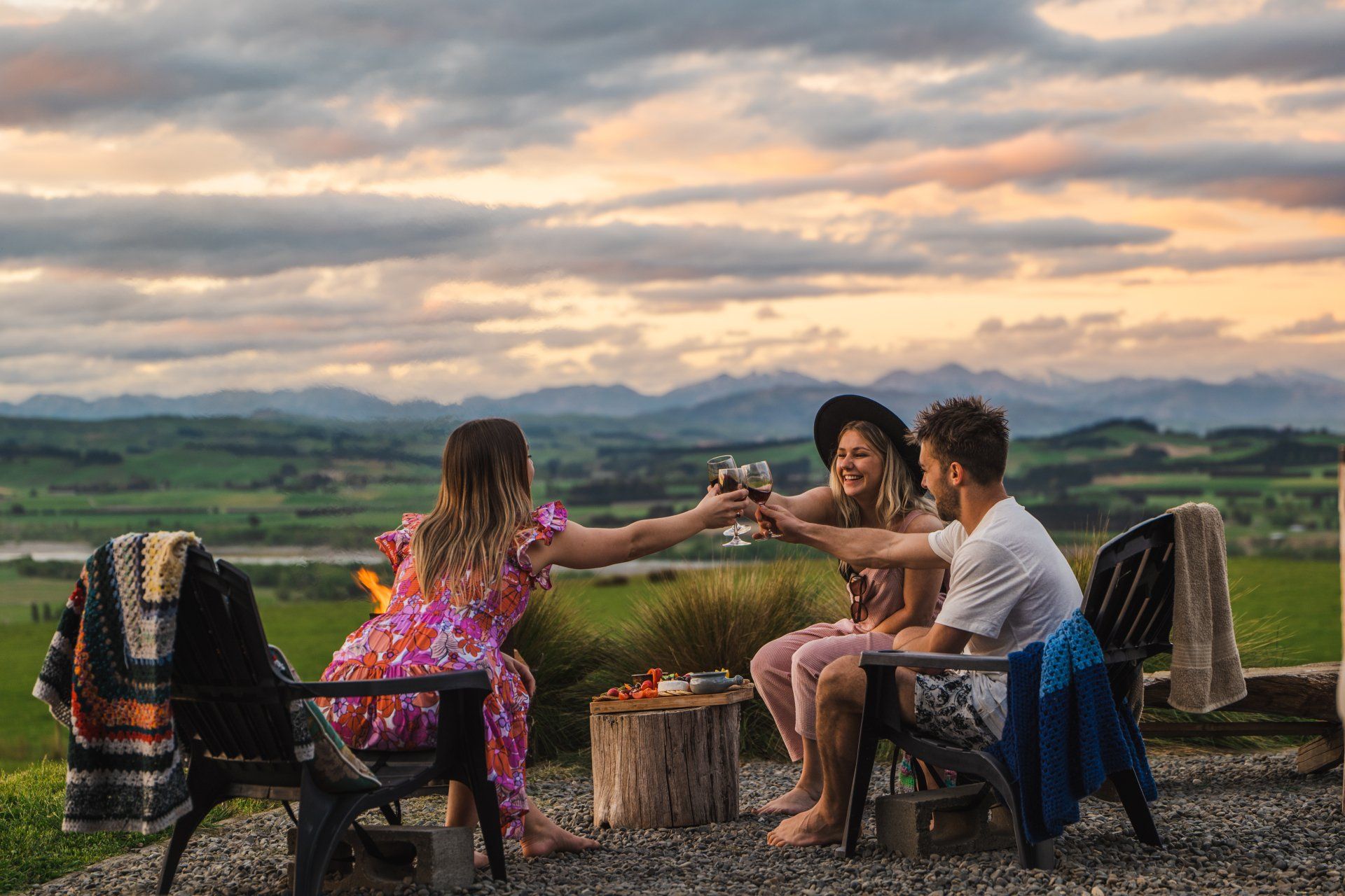 A group of people are sitting around a fire pit toasting with wine glasses