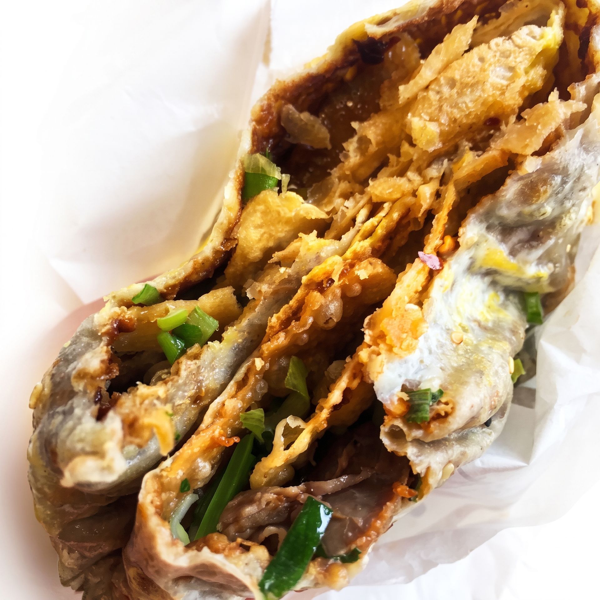 A crispy stuffed sandwich with fried filling, green onions, and dark sauce wrapped in white paper.