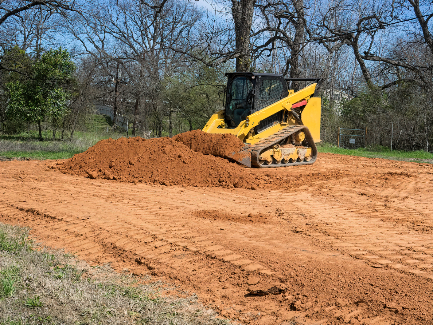 Yellow track loader moving a pile of reddish-brown dirt on a cleared area, trees in the background.