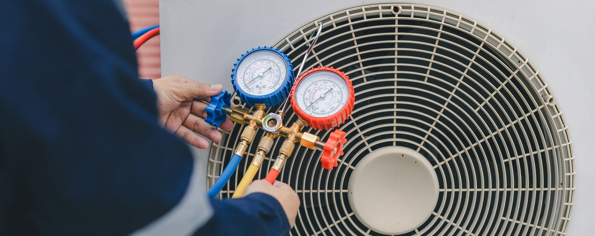HVAC technician connecting gauges to an AC unit. Blue and red gauges, a fan, and blue clothing are visible.