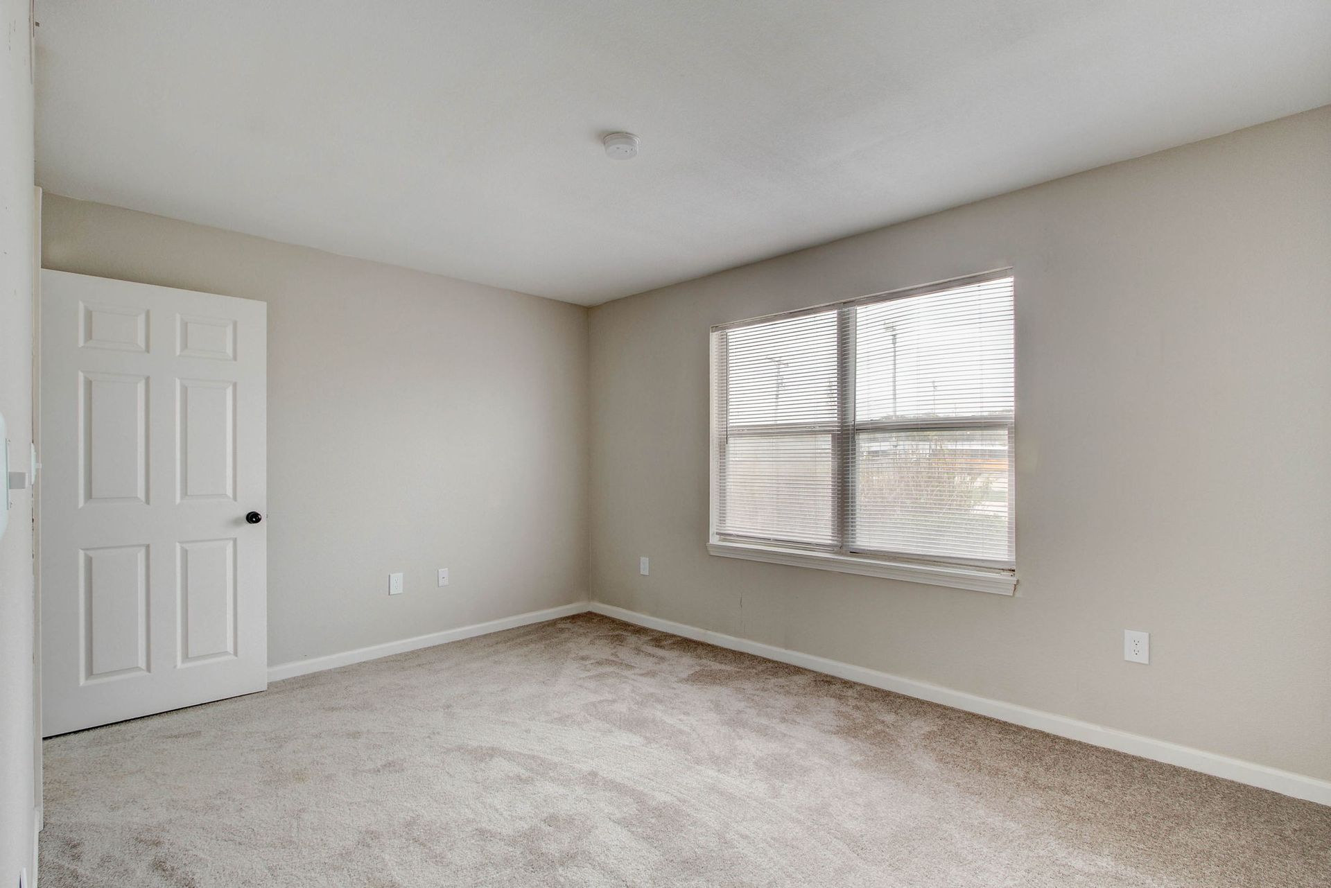 Empty bedroom with beige walls, white door, window with blinds, and light brown carpet.