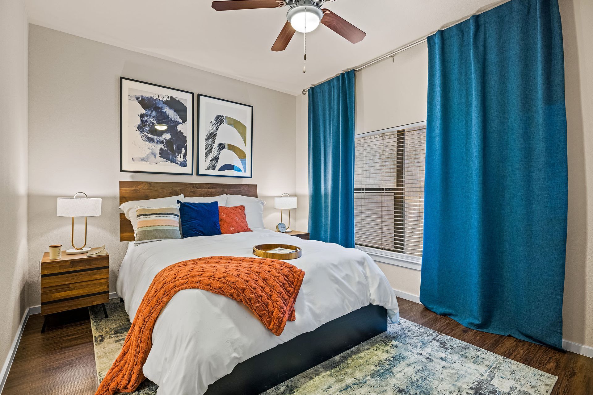 Apartment bedroom with a bed, nightstand, ceiling fan, and blue curtains at Marquis SoCo in South Austin, TX.
