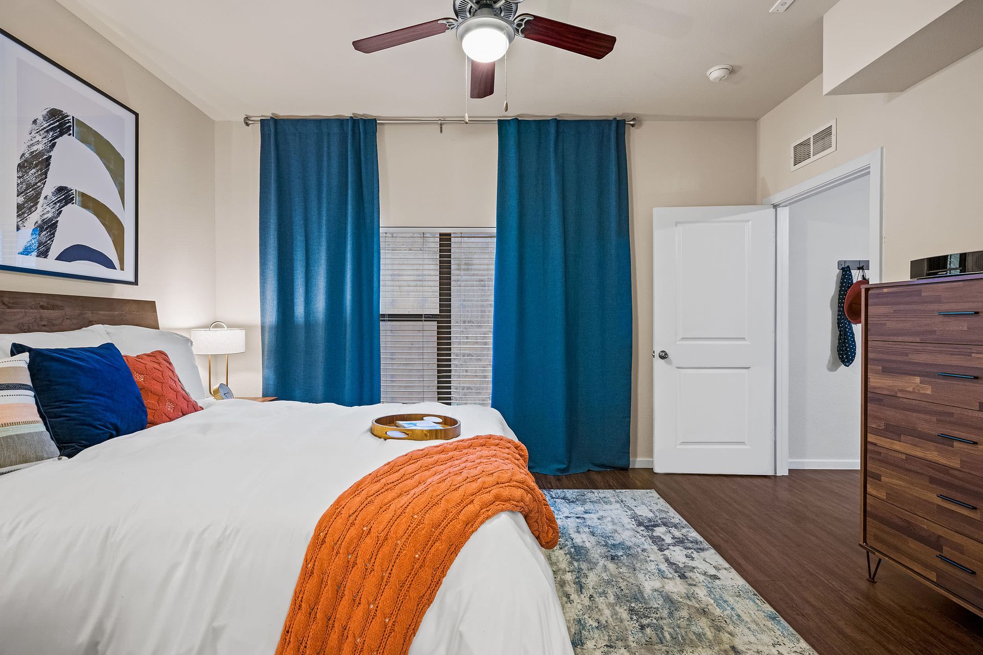 Apartment bedroom with a bed, dresser, ceiling fan, and blue curtains at Marquis SoCo in South Austin, TX.