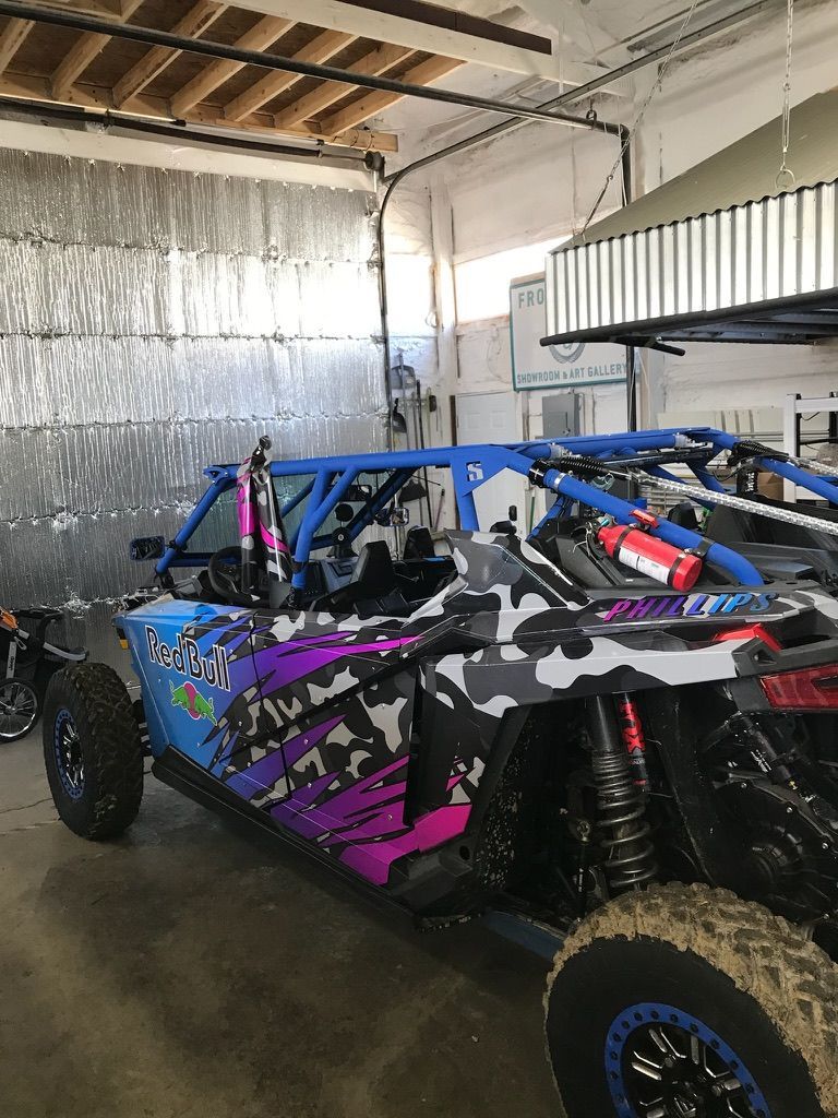 Colorful off-road buggy parked in a garage, with blue frame, black-purple wrap, and large tires.