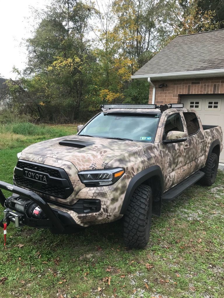 Camouflaged pickup truck parked beside a house on a grassy yard, with front winch and off-road tires.