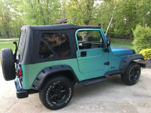 Teal Jeep Wrangler with black soft top parked on a driveway near trees