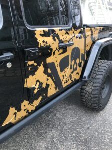 Black Jeep door with yellow and black splatter paint, parked on pavement with off-road tire visible