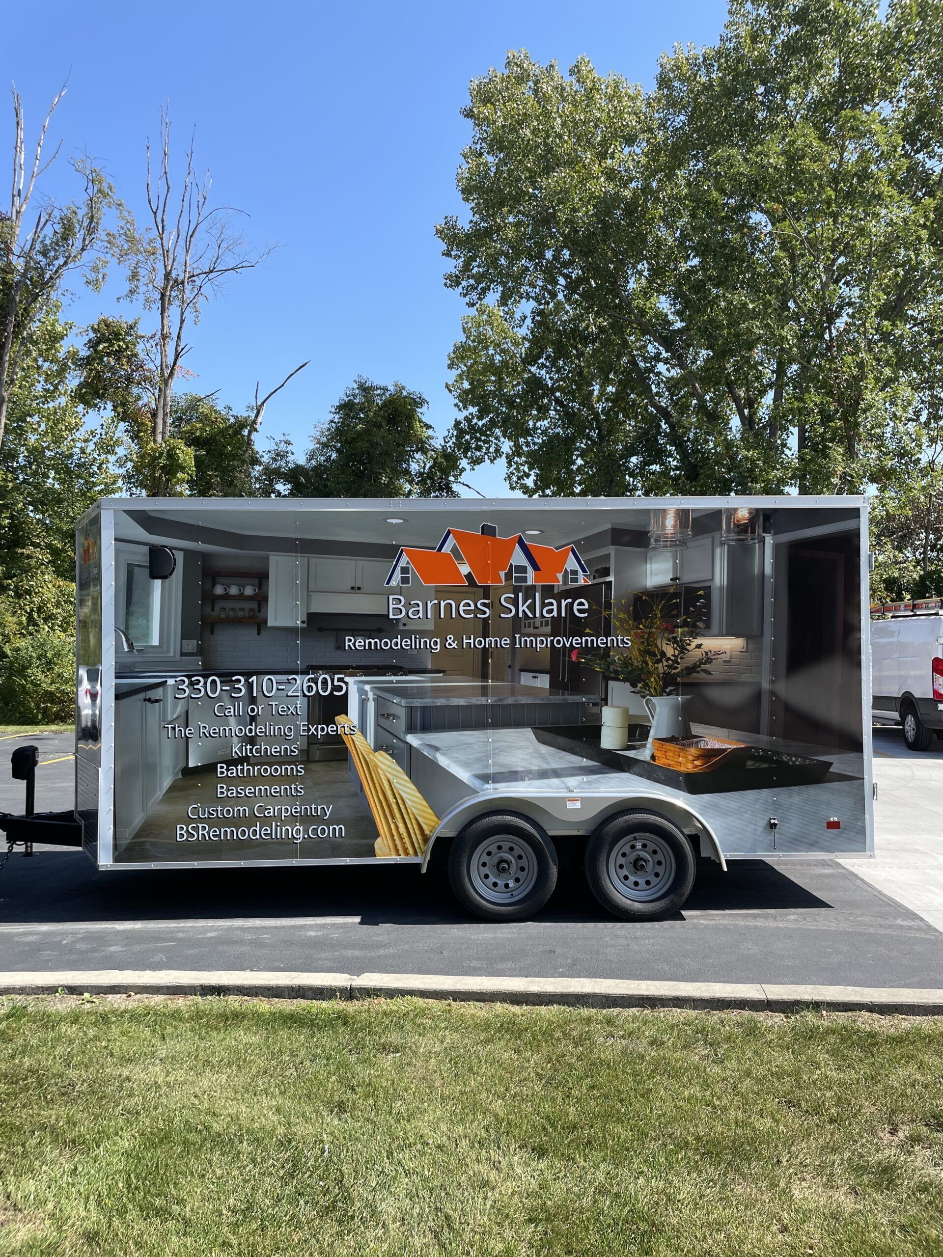 Silver food trailer parked outdoors on grass under trees, with orange logo and serving window.