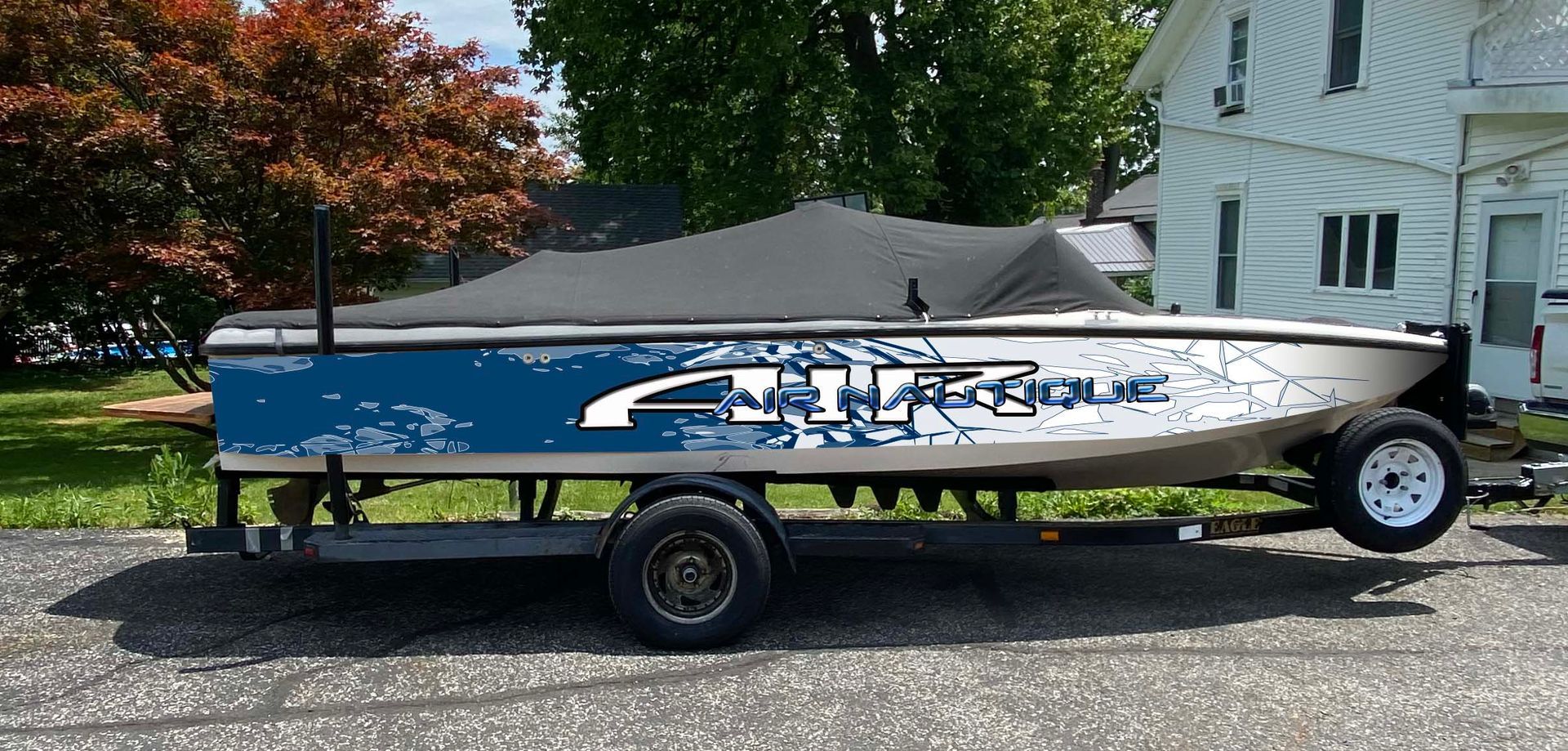 Small blue-and-silver boat on a trailer parked on a gravel driveway beside a house