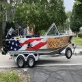 Small patriotic boat on a trailer with stars, stripes, and “We the People” lettering on a street.