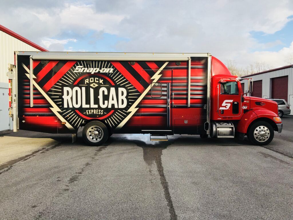 Red and black Rolling Rock “Roll Cab” delivery truck parked beside industrial buildings