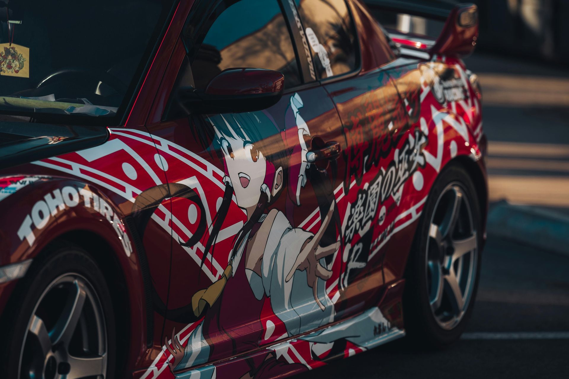 Red sports car with anime-style livery and decals, shown from the side in a garage.