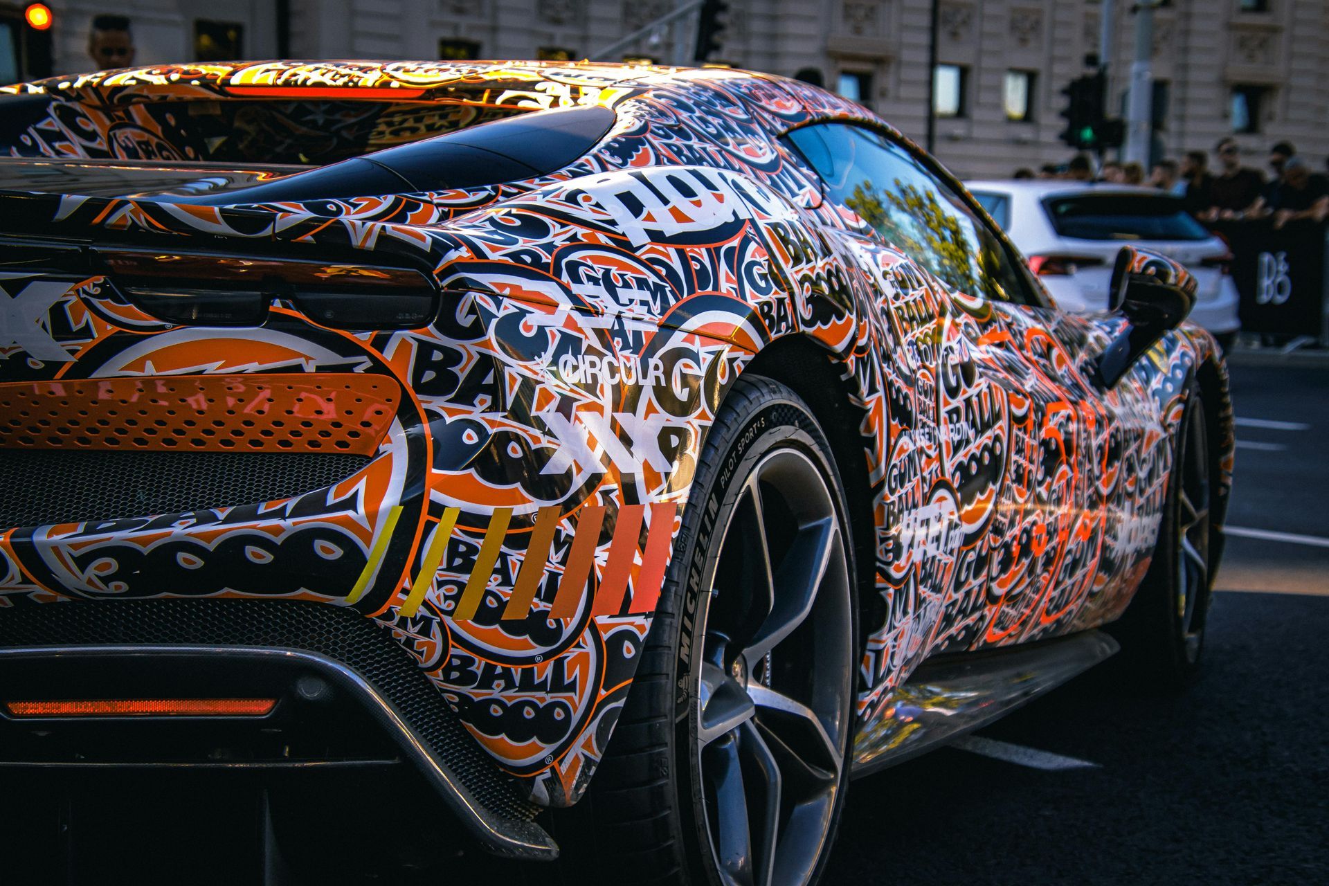 Camouflaged sports car wrapped in orange, black, and white graphics parked on a street at dusk