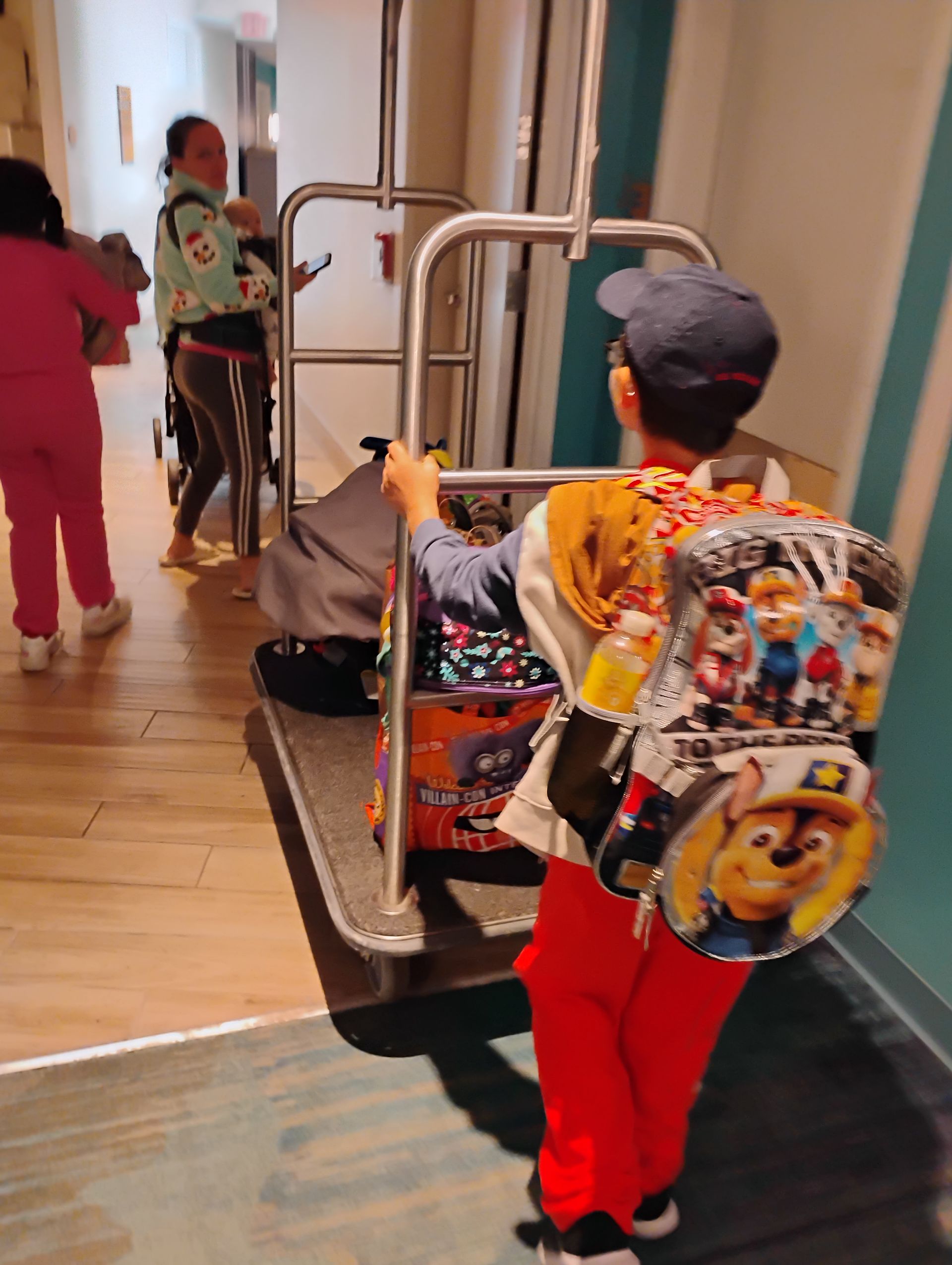 Boy pulling a luggage cart, wearing a Paw Patrol backpack, followed by family in a hotel hallway.