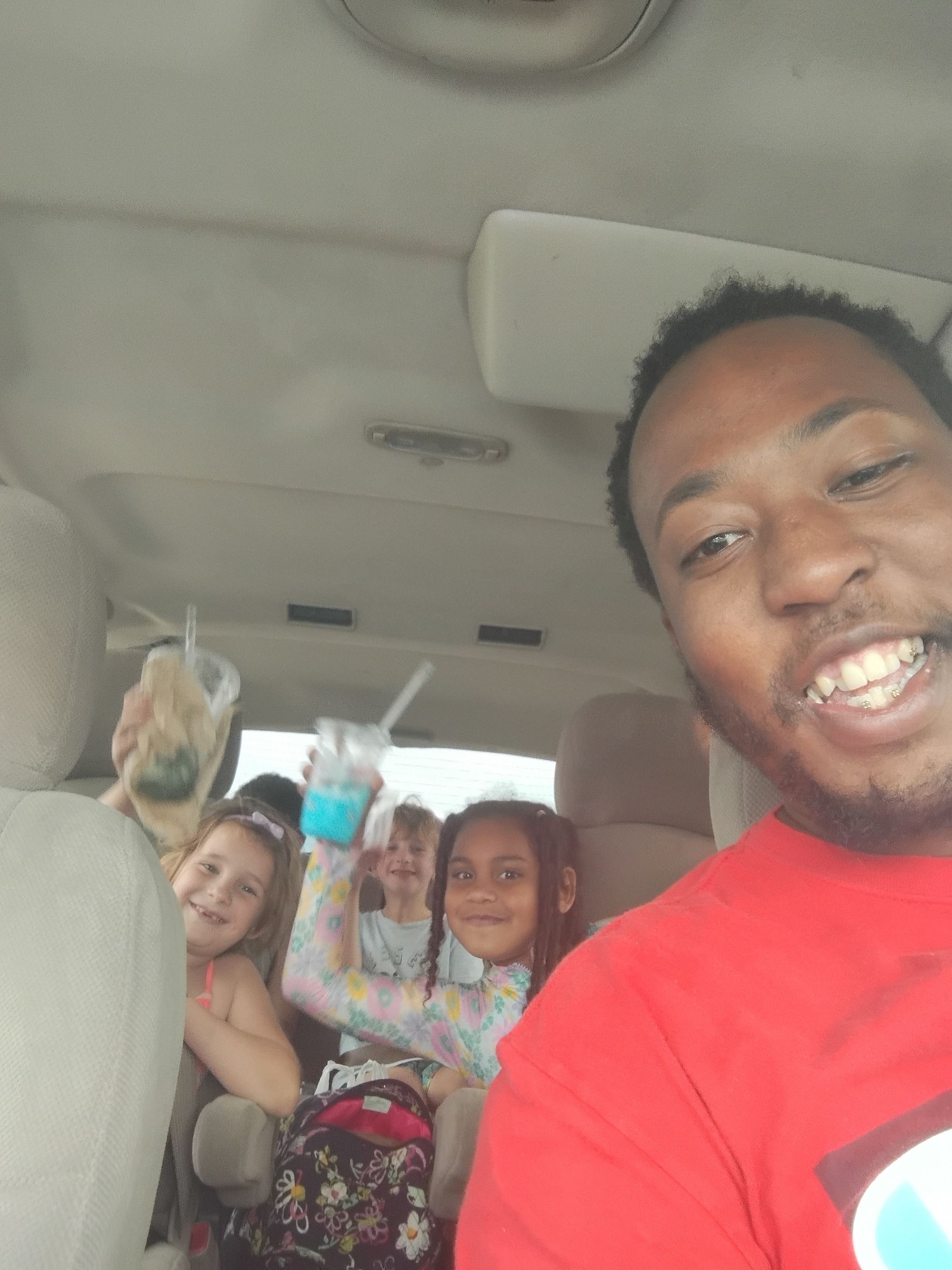 Man smiles for selfie in car with three young girls, one holding a blue drink.