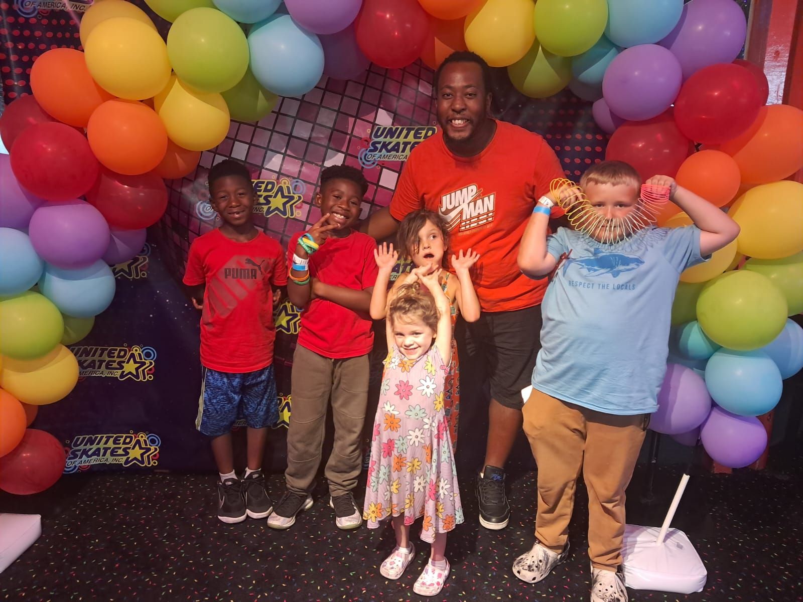 Group of children and an adult smiling in front of a balloon arch.