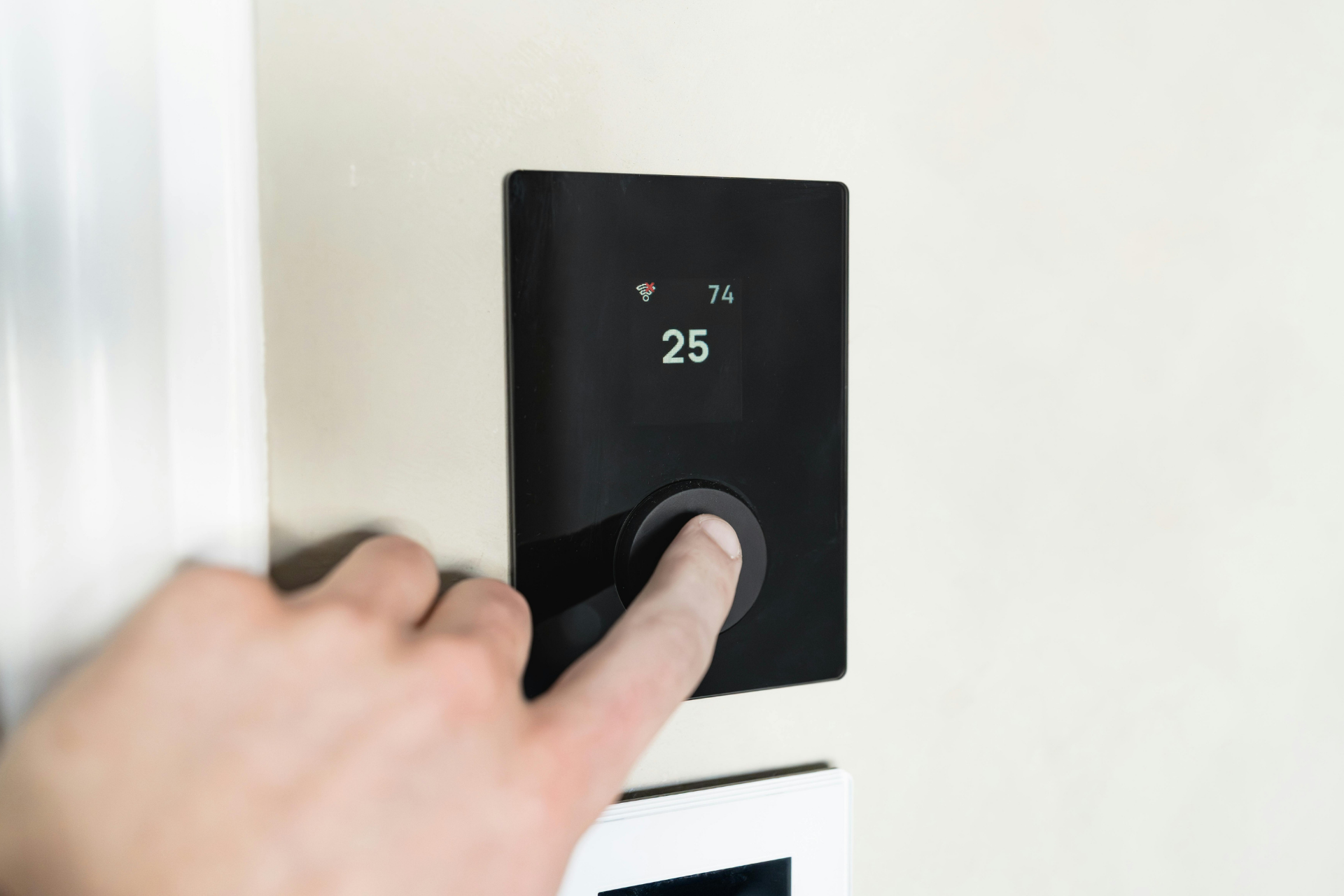 Hand adjusting a black wall thermostat showing 25°C on a white wall