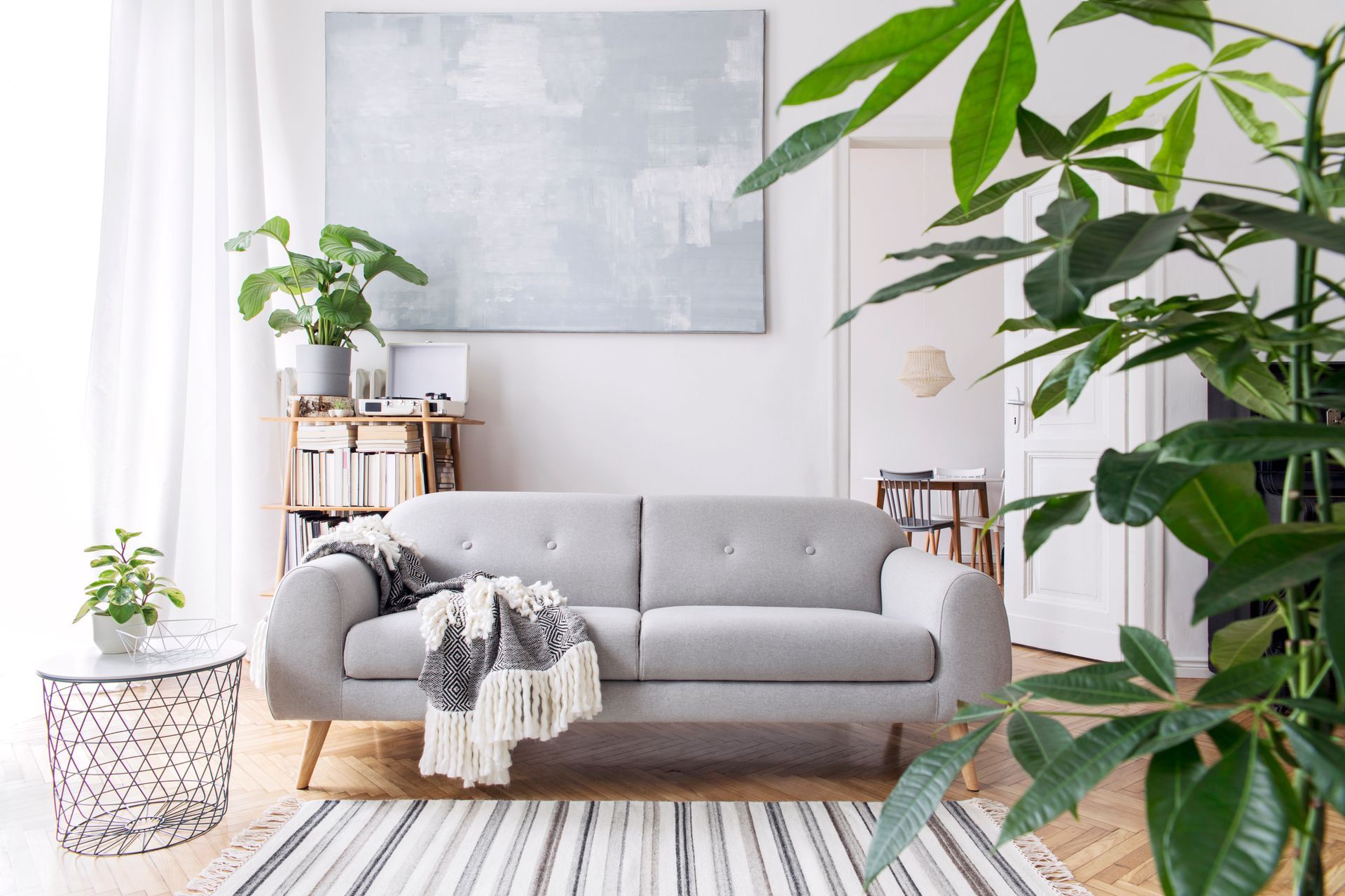A living room with a couch , rug , plants and a painting on the wall.