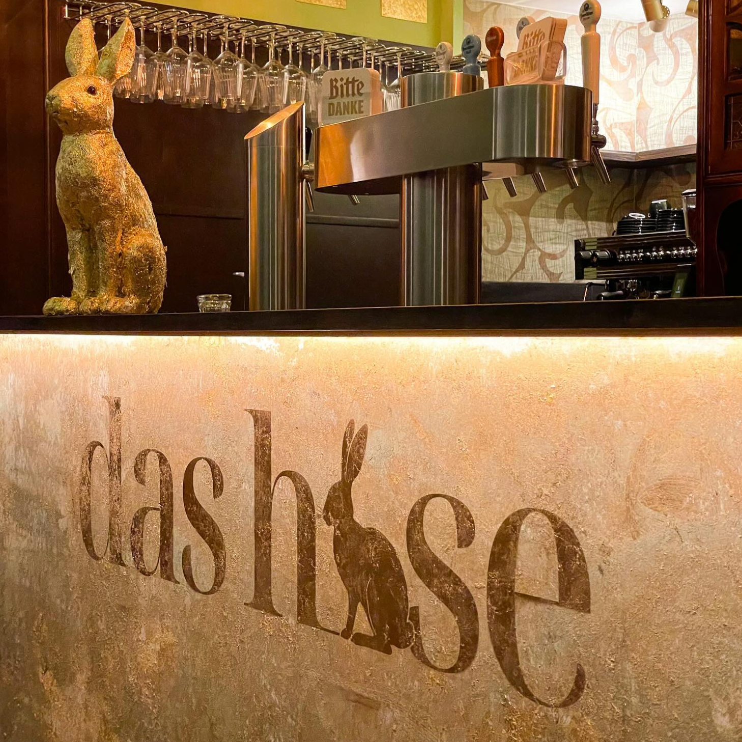 Restaurant-Empfangstresen mit hängenden Weingläsern und einer dekorativen Katzenstatue; Schild mit der Aufschrift „das hase“.