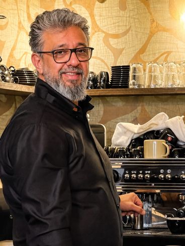 Ein Mann steht hinter einem Café-Tresen mit Espressomaschine und gestapelten Tassen und lächelt in die Kamera.