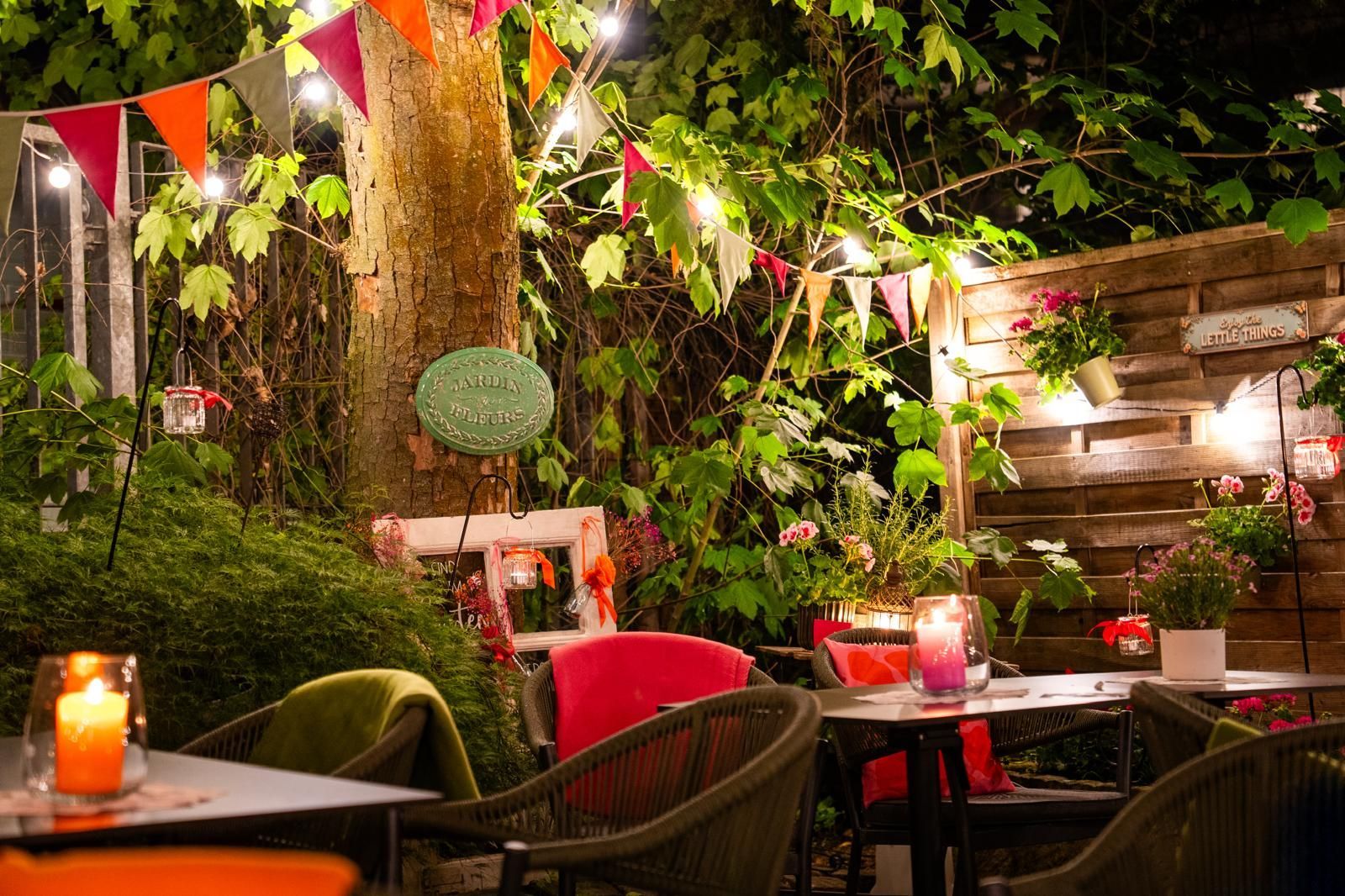 Gemütliche Außenterrasse eines Cafés bei Abend mit Lichterketten, Kerzen und Grünpflanzen um Tische und Stühle