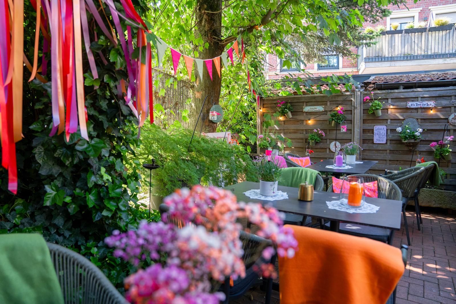 Farbenfrohe Gartencafé-Terrasse mit Girlanden, Topfblumen und Tischen unter Bäumen