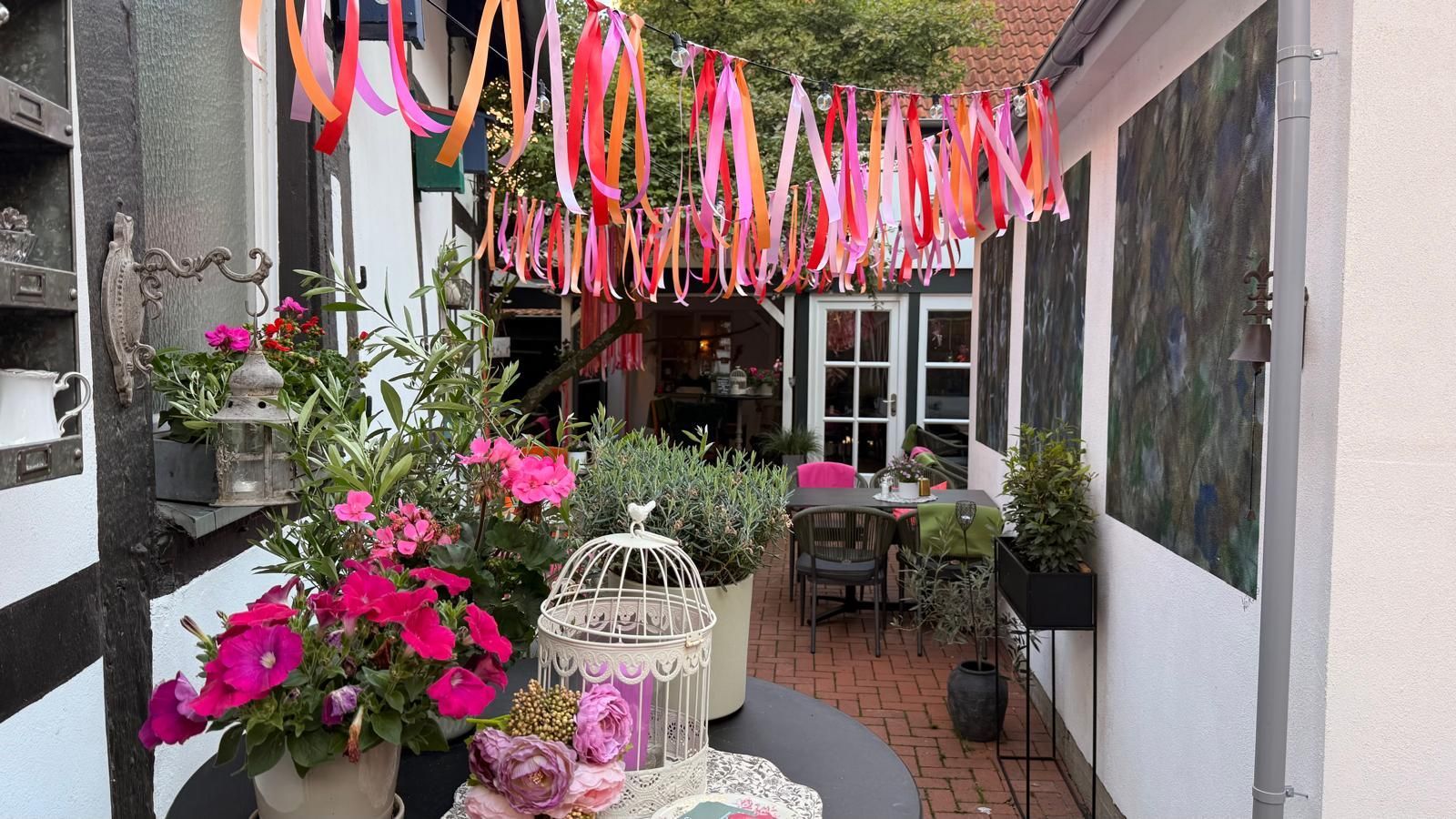 Ein mit bunten Bändern geschmückter Innenhof mit rosa Blumen, weißen Stühlen und einem gemütlichen, caféähnlichen Sitzbereich